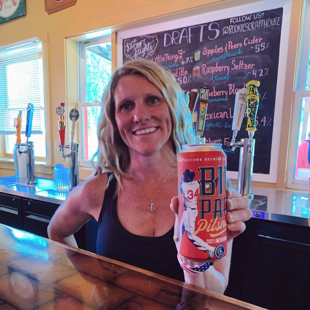 Woman behind the bar holding a can of beer, with the draft list on a chalkboard behind her