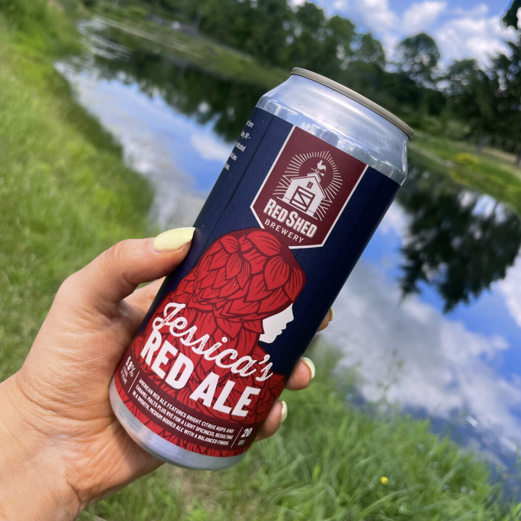 Hand holding a can of Red Shed Jessica's Red Ale by the water